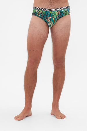 Crop view of model wearing Hotel Franks by CAMILLA mens green swim briefs in Sing My Song Print