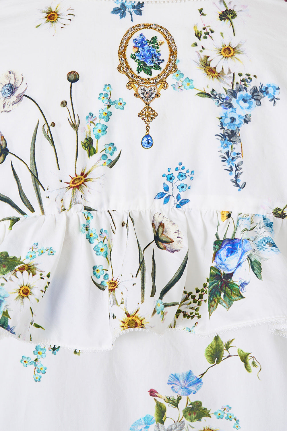 White tiered dress featuring a colorful floral print with blue flowers, green leaves, and a gold and blue brooch motif printed on the bodice.